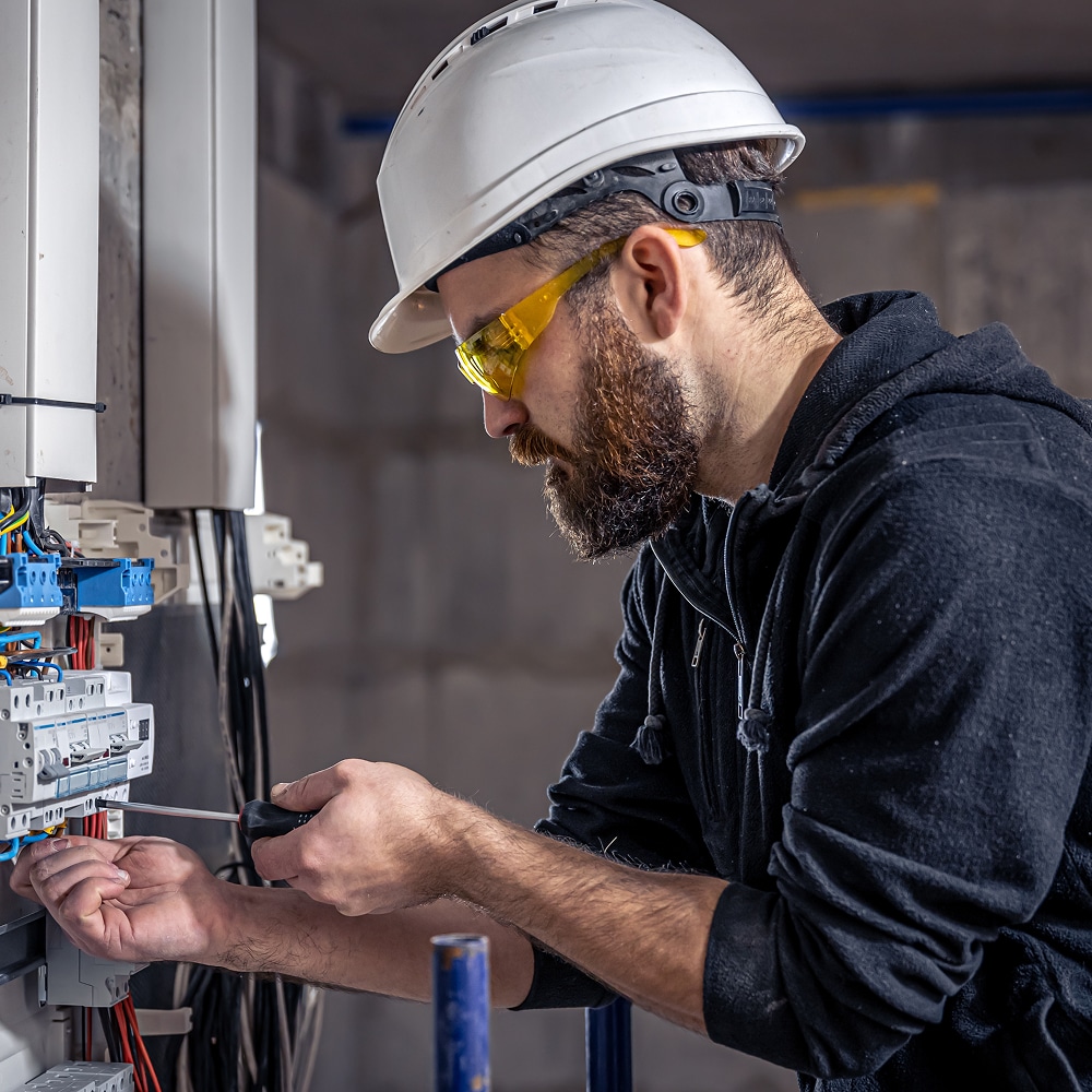 Électricien barbu avec casque et lunettes travaillant sur un tableau électrique au tournevis.